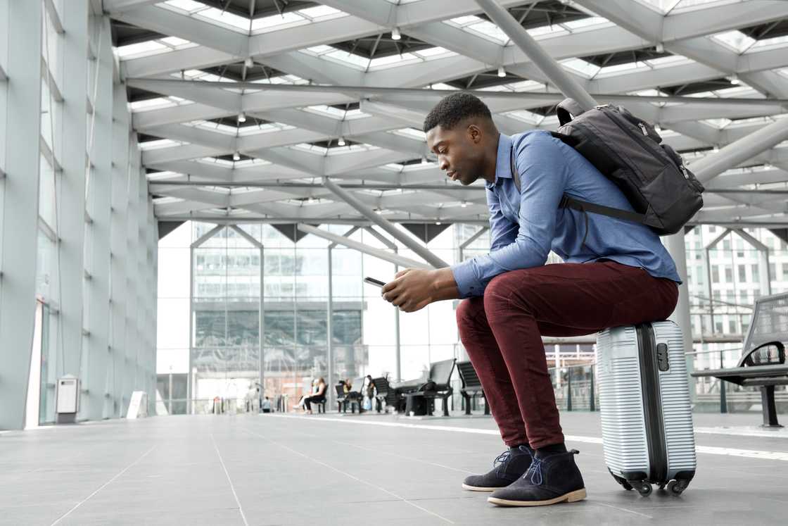 Person sits on a silver suitcase in a modern terminal, looking at a smartphone with a backpack on. Person sits on a silver suitcase in a modern terminal, looking at a smartphone with a backpack on.