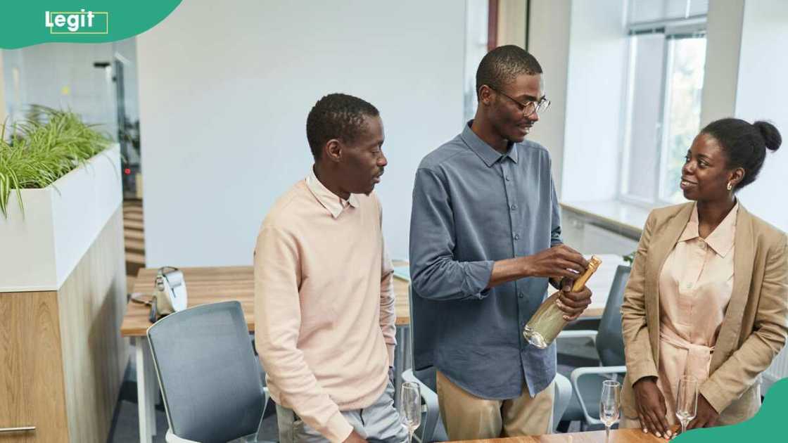A man opening a bottle of wine as his tow colleagues look on A man opening a bottle of wine as his tow colleagues look on