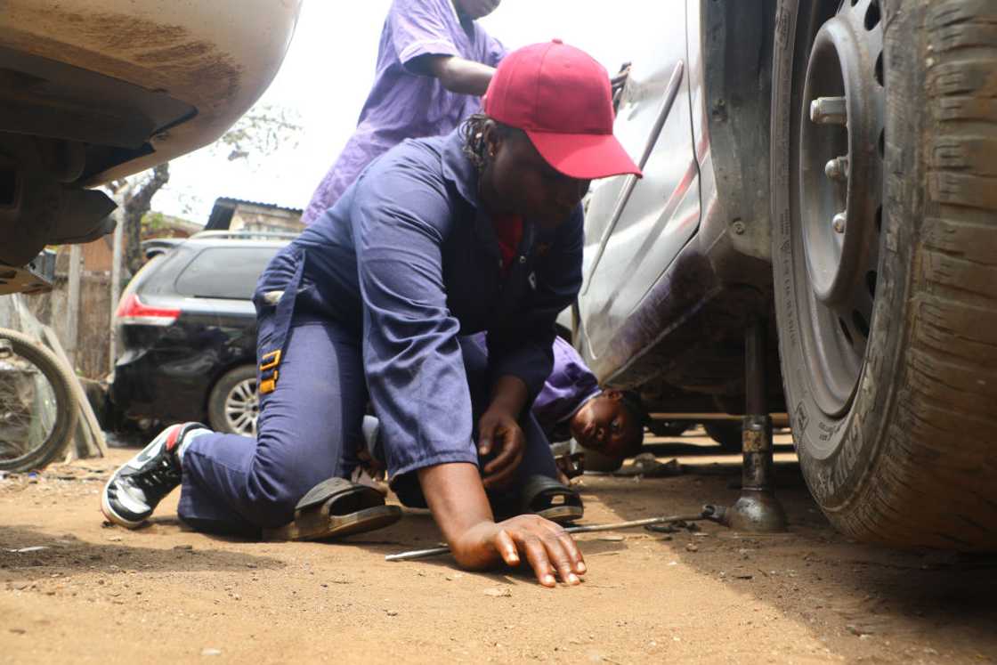 Mechanical engineer Ajaye Oluwatosin in navy overalls and a red cap, kneeling beside a raised car as she works on it at her workshop in Lagos, 2023