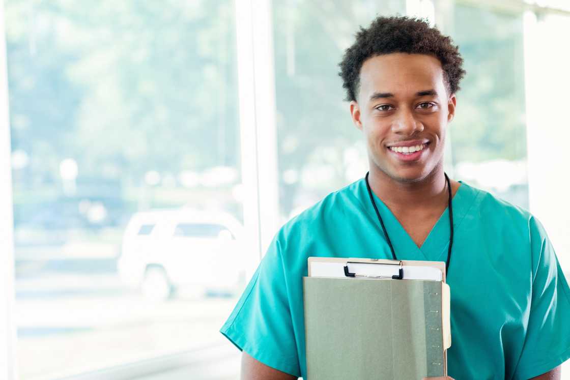 A young man in teal scrubs holds a folder and clipboard, smiling at the camera in front of large windows.