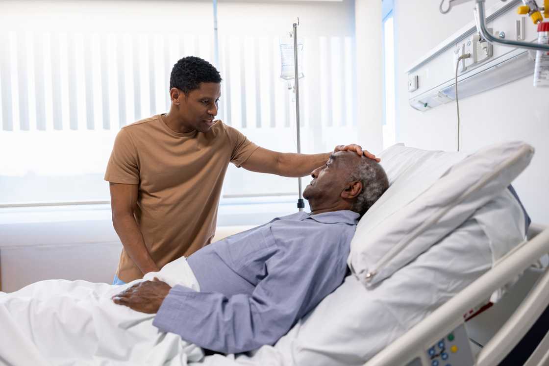 Younger person touches forehead of elderly patient in hospital bed.