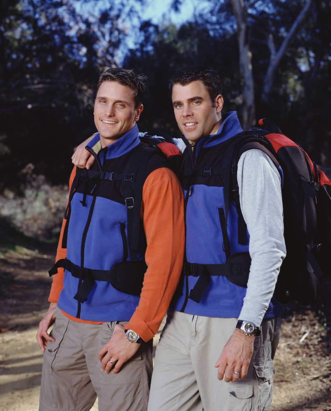Two men in matching blue fleece vests and backpacks stand close together, smiling, in a wooded outdoor setting.