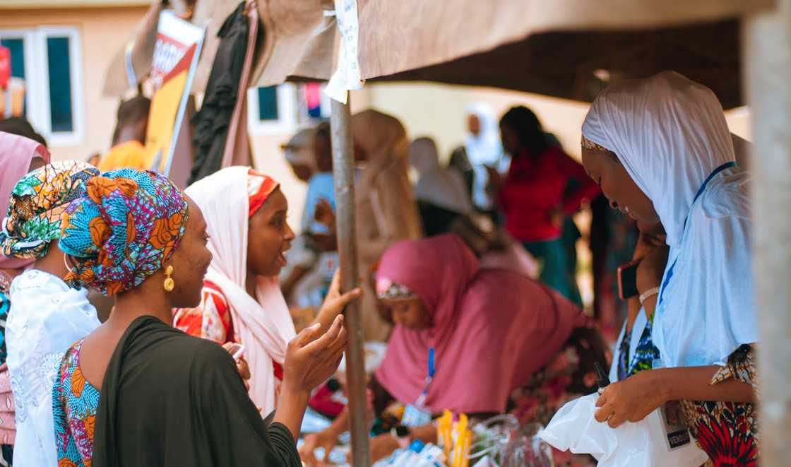 Several women wearing headscarves talk and trade items at a busy outdoor market.