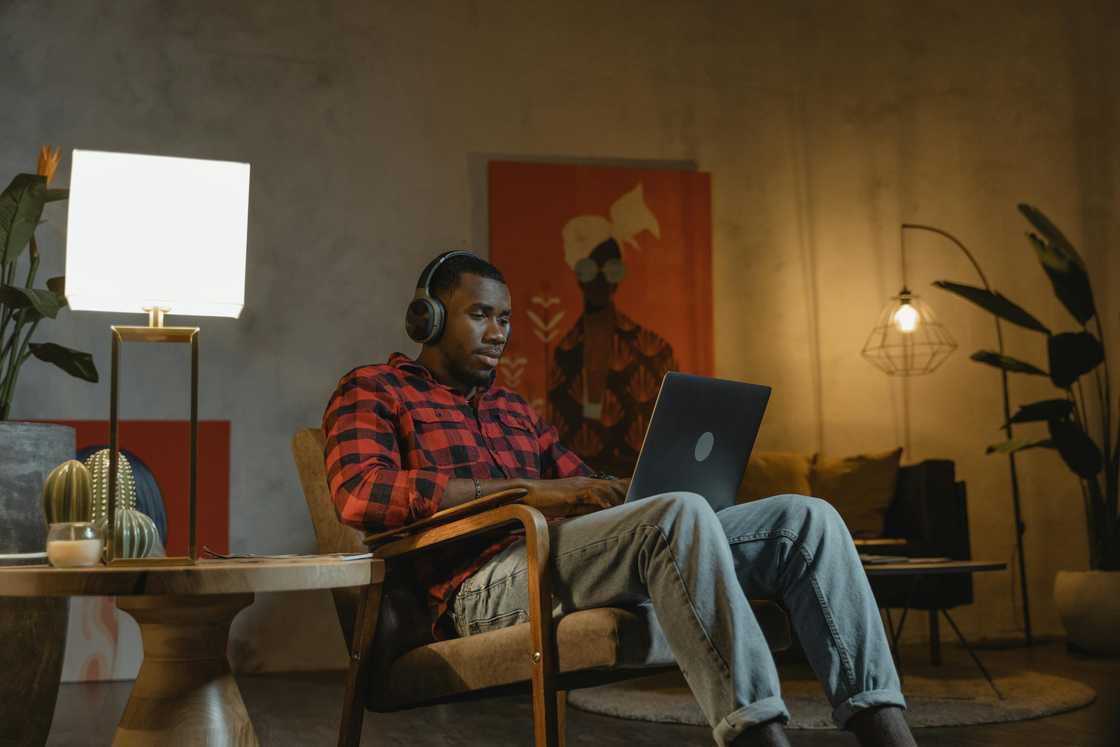 A man in a checkered shirt and denim pants is pictured using a laptop