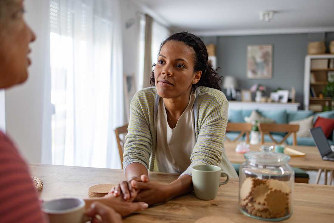 Two people sit at a wooden table; one speaks while the other listens and holds their hand. Two people sit at a wooden table; one speaks while the other listens and holds their hand.