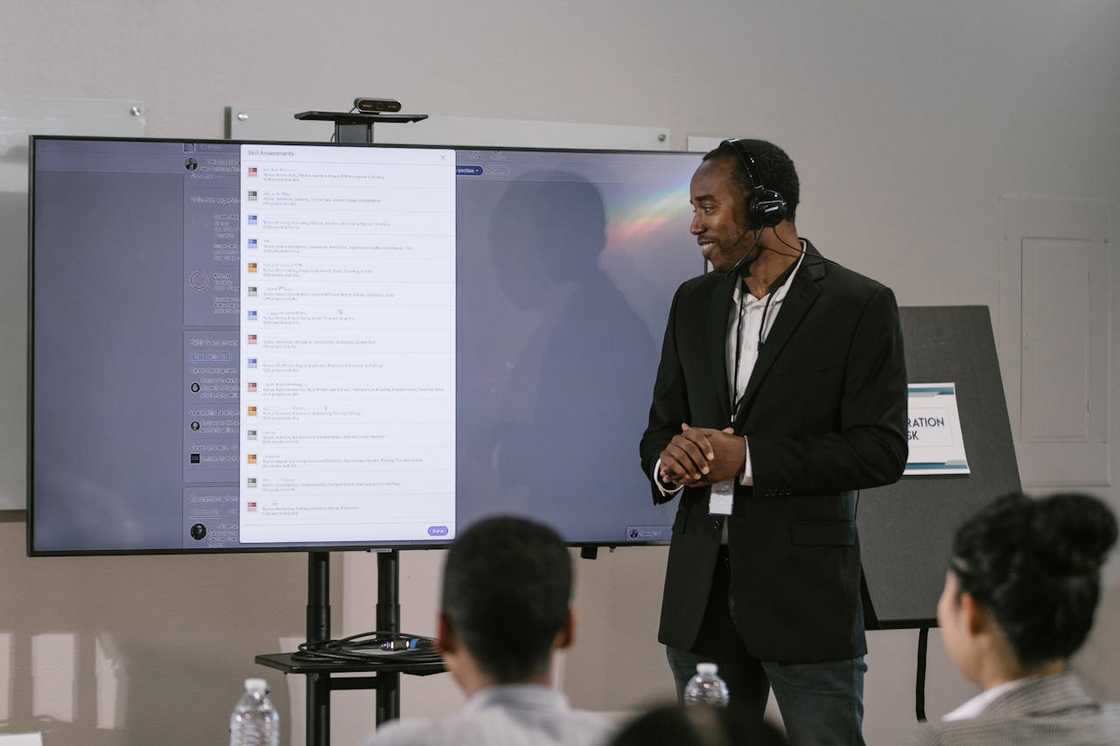 A man wearing a headset presents information on a large screen to a seated audience. A man wearing a headset presents information on a large screen to a seated audience.