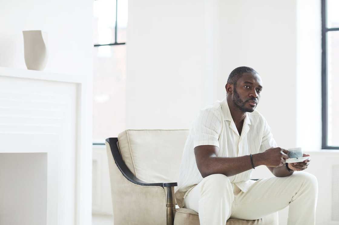 A man sits in an armchair holding a cup in a bright room.