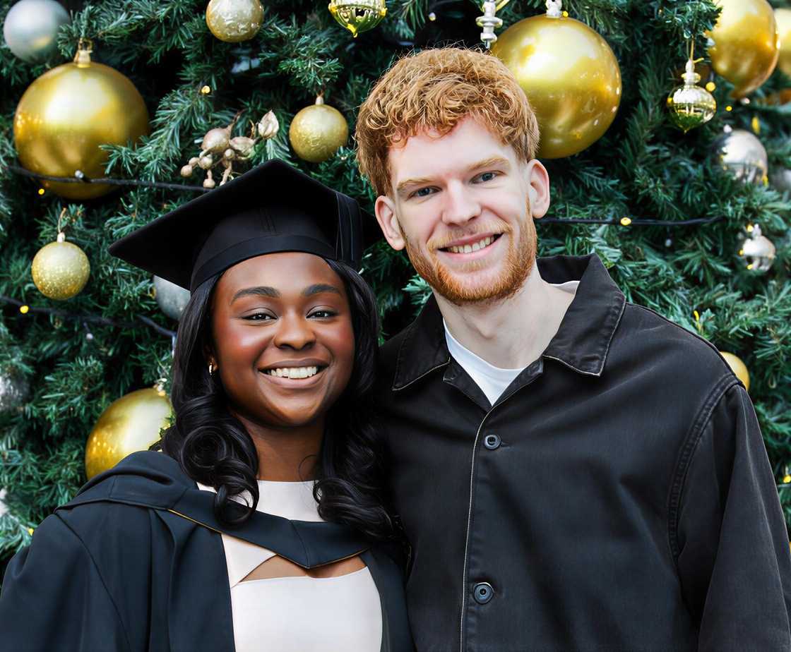 A female student wearing a graduation gown and male students posing next to a Christmas tree A female student wearing a graduation gown and male students posing next to a Christmas tree
