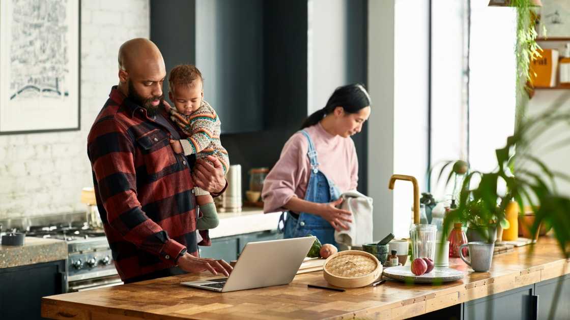 A man in a red and black shirt holds a baby as he works on a laptop, while a woman in pink and blue clothes holds a utensil. A man in a red and black shirt holds a baby as he works on a laptop, while a woman in pink and blue clothes holds a utensil.