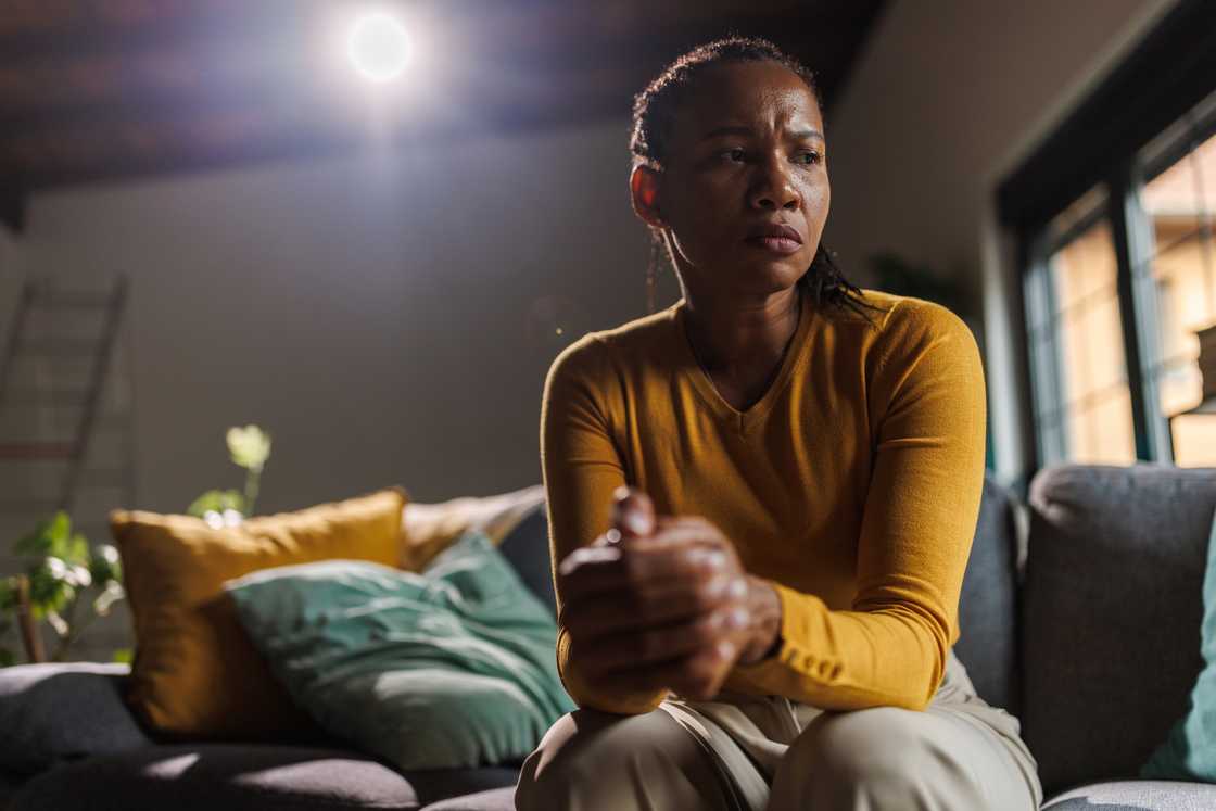 A woman in a mustard yellow shirt sits on a couch, hands clasped, in a softly lit room. A woman in a mustard yellow shirt sits on a couch, hands clasped, in a softly lit room.