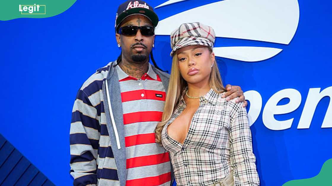 21 Savage in an Atlanta cap and layered striped outfit stands with a woman in a plaid shirt in front of a US Open backdrop. 21 Savage in an Atlanta cap and layered striped outfit stands with a woman in a plaid shirt in front of a US Open backdrop.