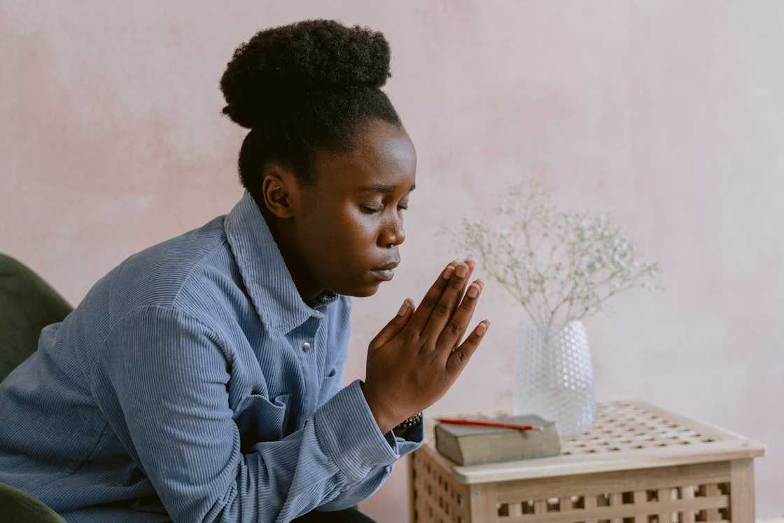 A woman with hands clasped in prayer, leaning forward beside a small table with a book. A woman with hands clasped in prayer, leaning forward beside a small table with a book.