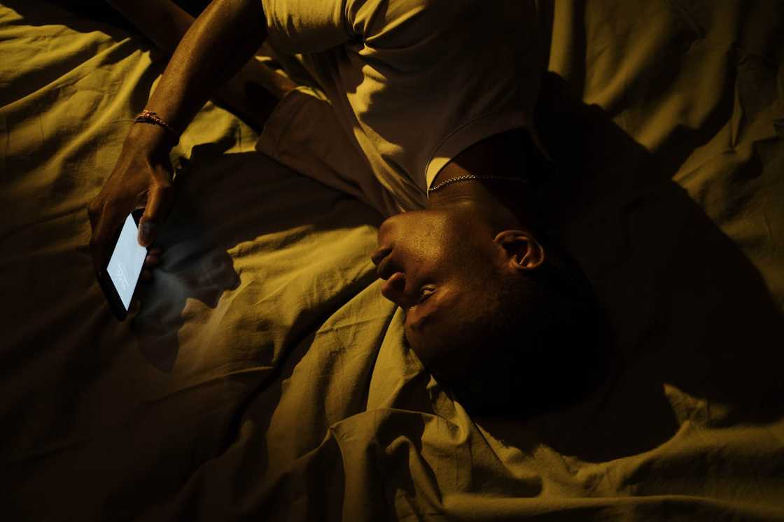 A man lies on a bed holding a phone in dim light.