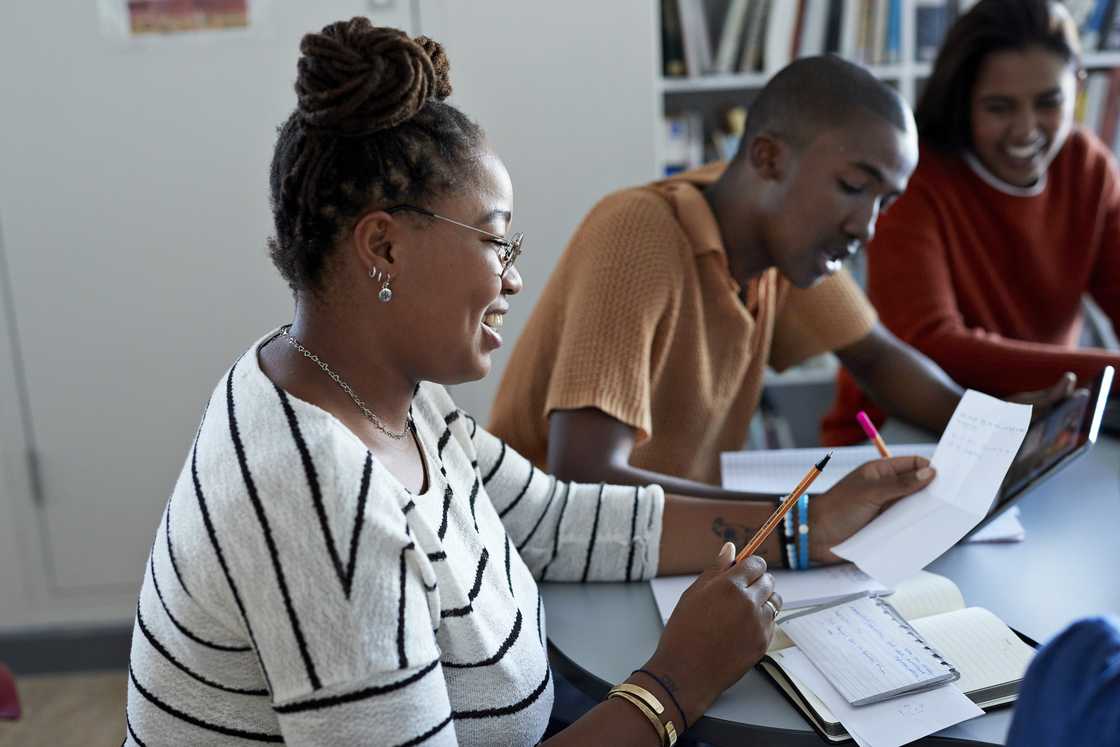 Side view of smiling young female student reading paper while sitting by friends at desk in library.