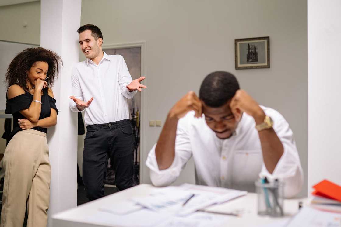 A man sits at a desk holding his head while two people stand behind him gossiping.