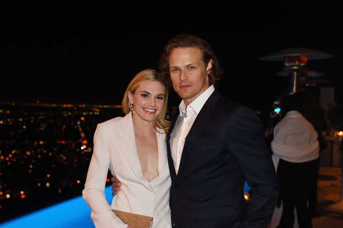 Mackenzie Mauzy in a white outfit (L) and Sam Heughan in a black suite and a white shirt (R) posing for a photo Mackenzie Mauzy in a white outfit (L) and Sam Heughan in a black suite and a white shirt (R) posing for a photo