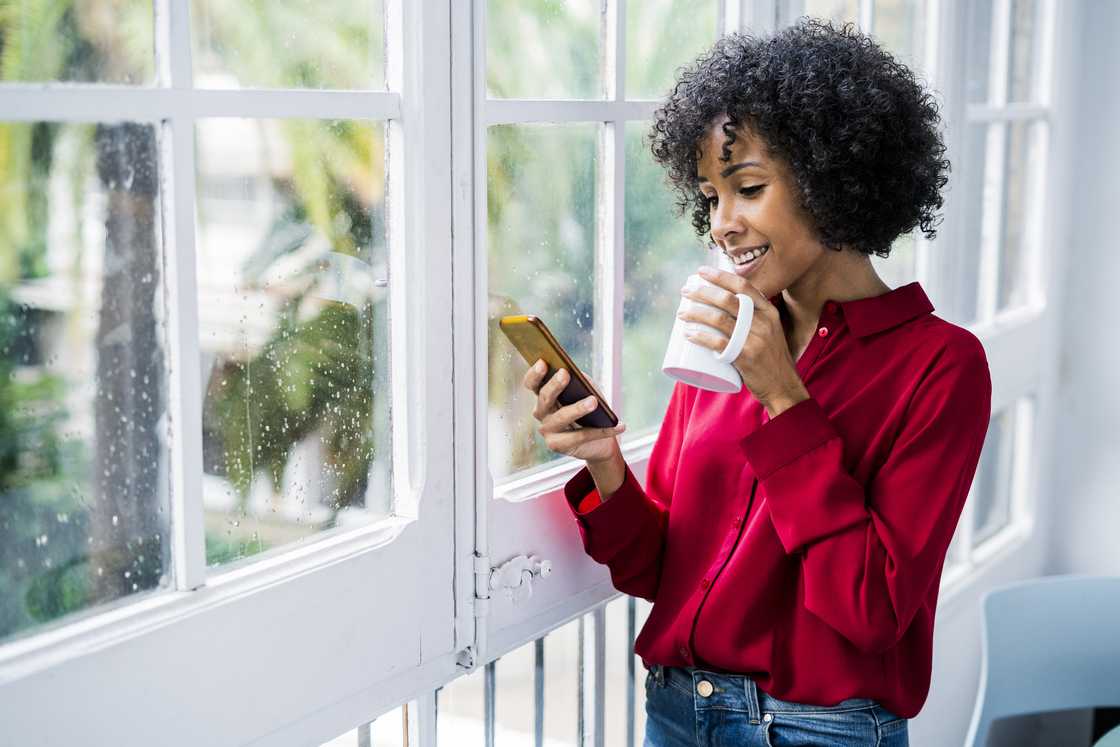 Smiling woman with cup of coffee and cell phone standing at the window at home