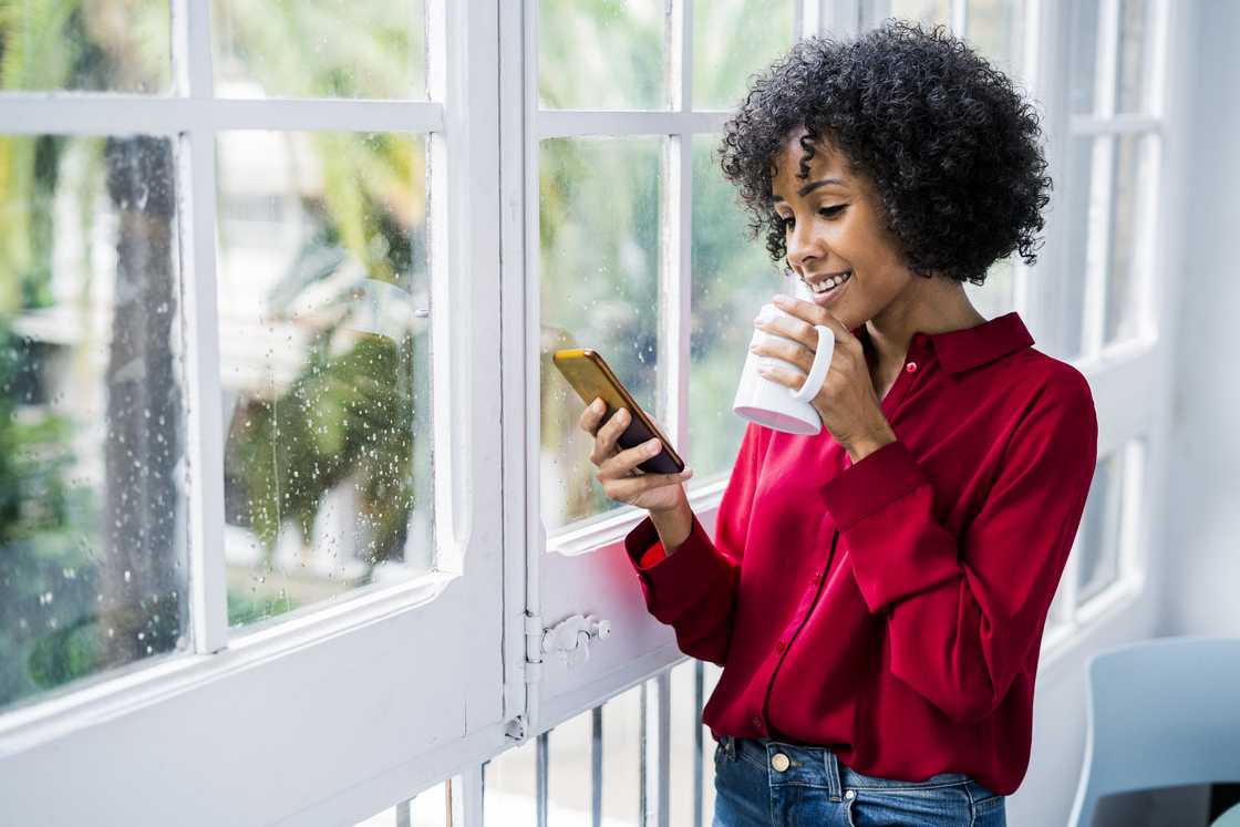 Smiling woman with cup of coffee and cell phone standing at the window at home Smiling woman with cup of coffee and cell phone standing at the window at home