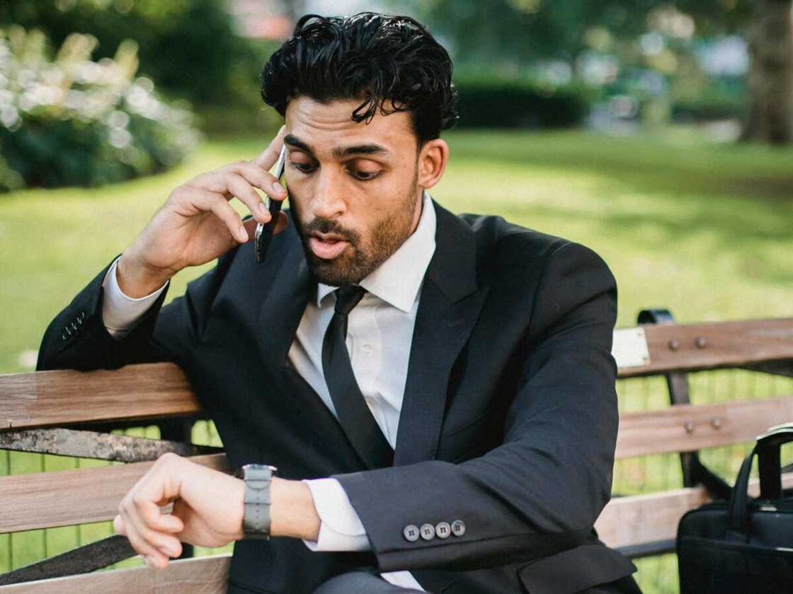 A man in a black suit sitting on a bench talking on the phone and looking at his watch