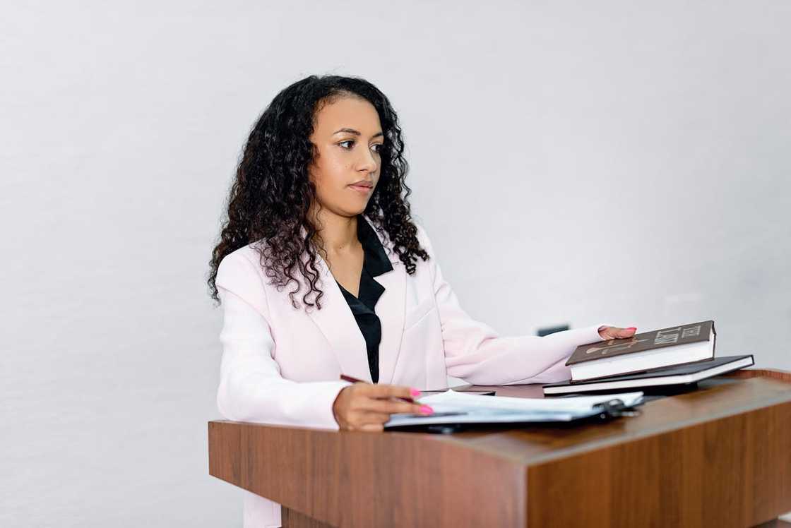 A woman in a pink blazer standing at a podium with books and documents.