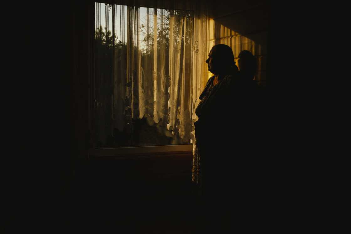 Silhouetted person stands near a sunlit window in a dimly lit room. Silhouetted person stands near a sunlit window in a dimly lit room.