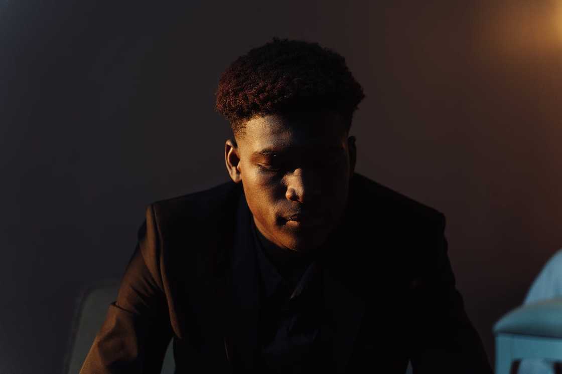 A man sits in low light with his head bowed, partially lit from the side. A man sits in low light with his head bowed, partially lit from the side.