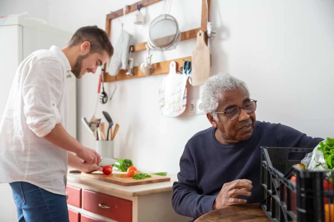 A young man chops vegetables while an elderly man sits nearby in the kitchen. A young man chops vegetables while an elderly man sits nearby in the kitchen.