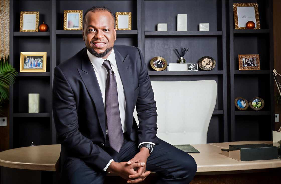 Igho Charles Sanomi poses in a black suit, sitting on a wooden desk in an office. Igho Charles Sanomi poses in a black suit, sitting on a wooden desk in an office.