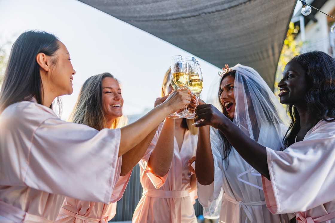 A joyful, diverse group of bridesmaids in matching robes cheer with the bride outdoors during a bachelorette celebration.