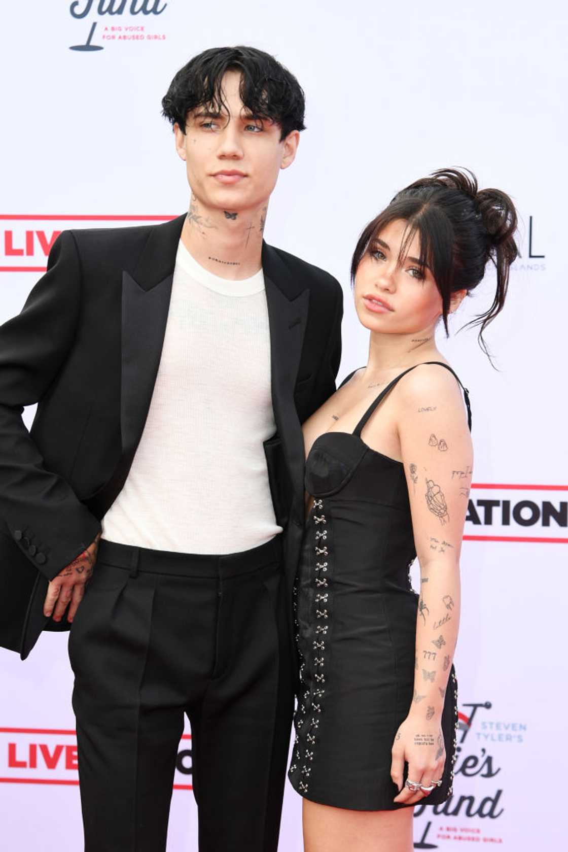 Jaden Hossler in a black suit over a white tee stands beside a woman in a black lace-up dress with visible tattoos. Jaden Hossler in a black suit over a white tee stands beside a woman in a black lace-up dress with visible tattoos.