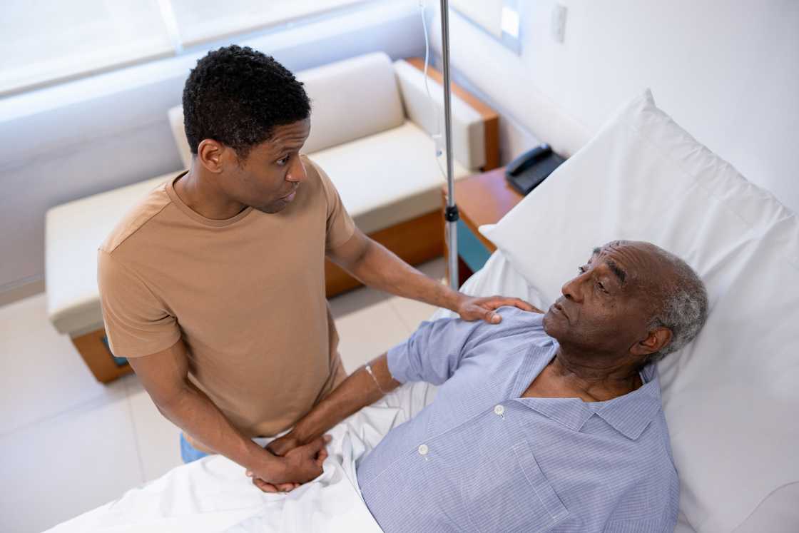 Younger person holds hands with older patient in hospital bed. Younger person holds hands with older patient in hospital bed.
