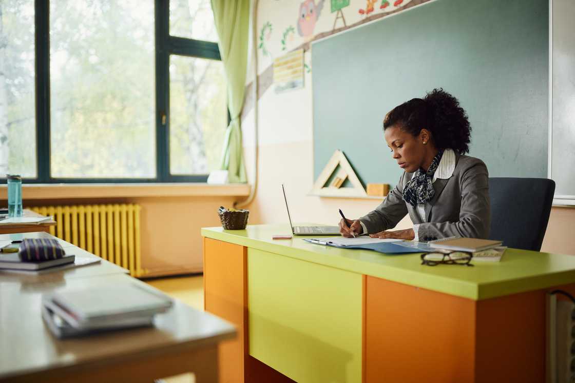 Teacher sits at a bright desk in a sunlit classroom, writing in a notebook.