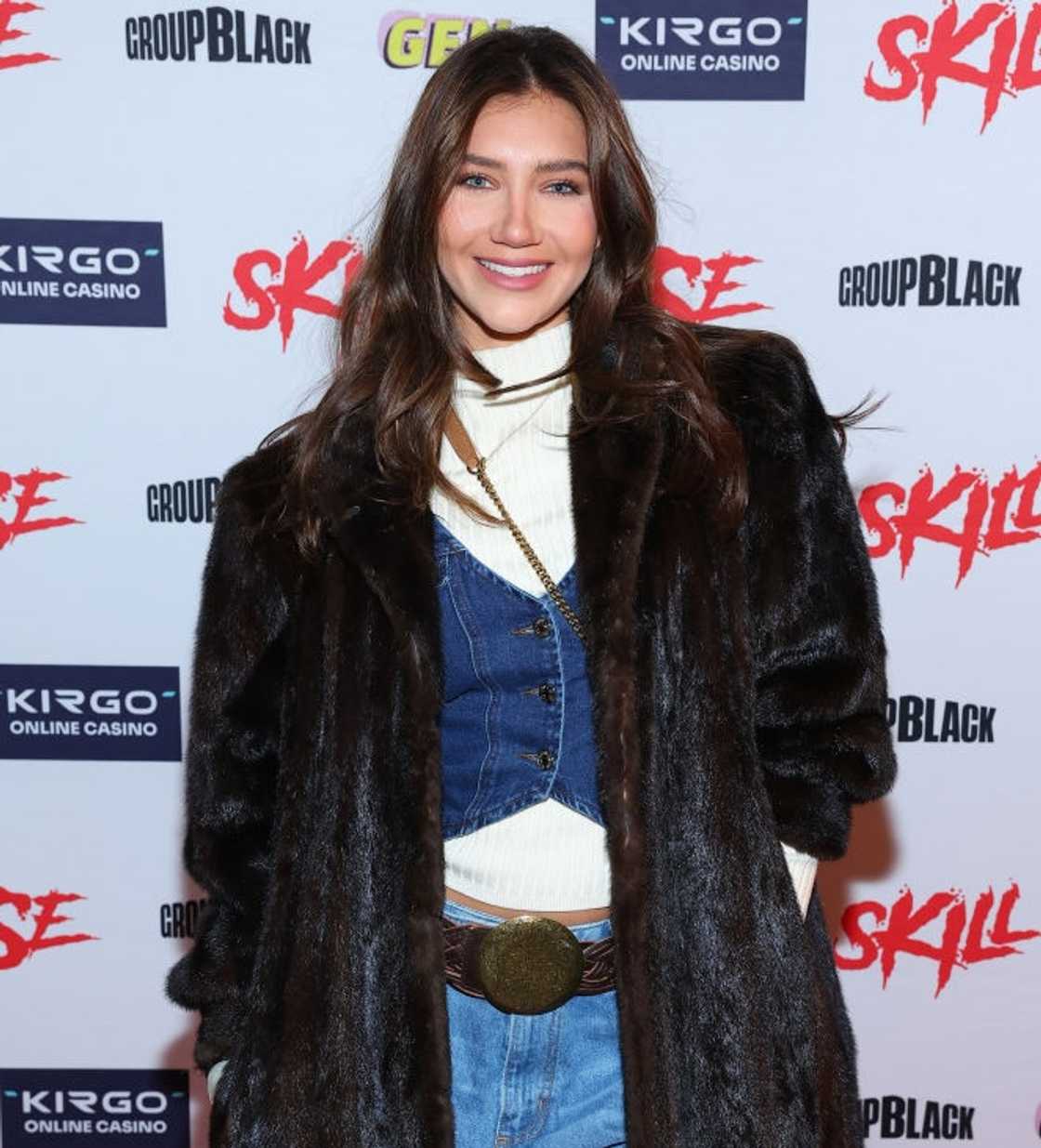 A woman in a long fur coat over a denim and white outfit poses in front of a Skillhouse step-and-repeat.