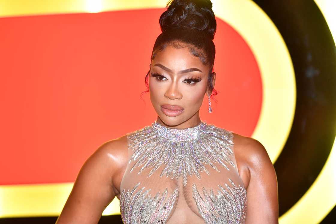 A woman with her hair in a high bun wears a sheer beaded gown in front of a red and yellow backdrop.
