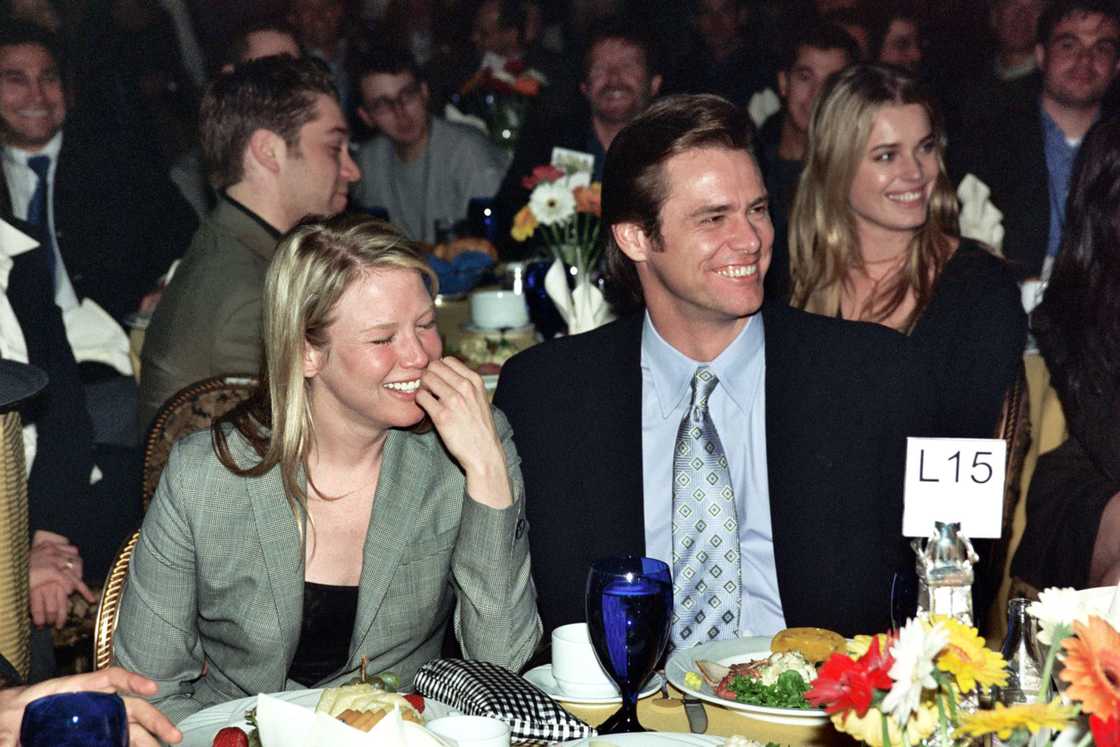 A young Jim Carrey in a suit and tie sits beside a laughing Renée Zellweger at a dinner table.