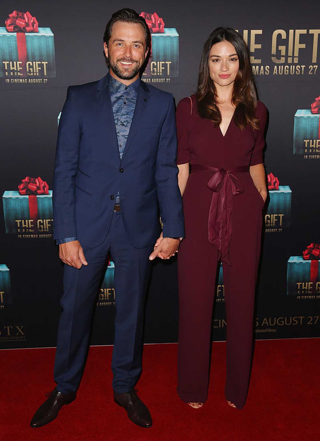 A bearded man in a navy suit holds hands with a woman in a burgundy jumpsuit at a step-and-repeat.