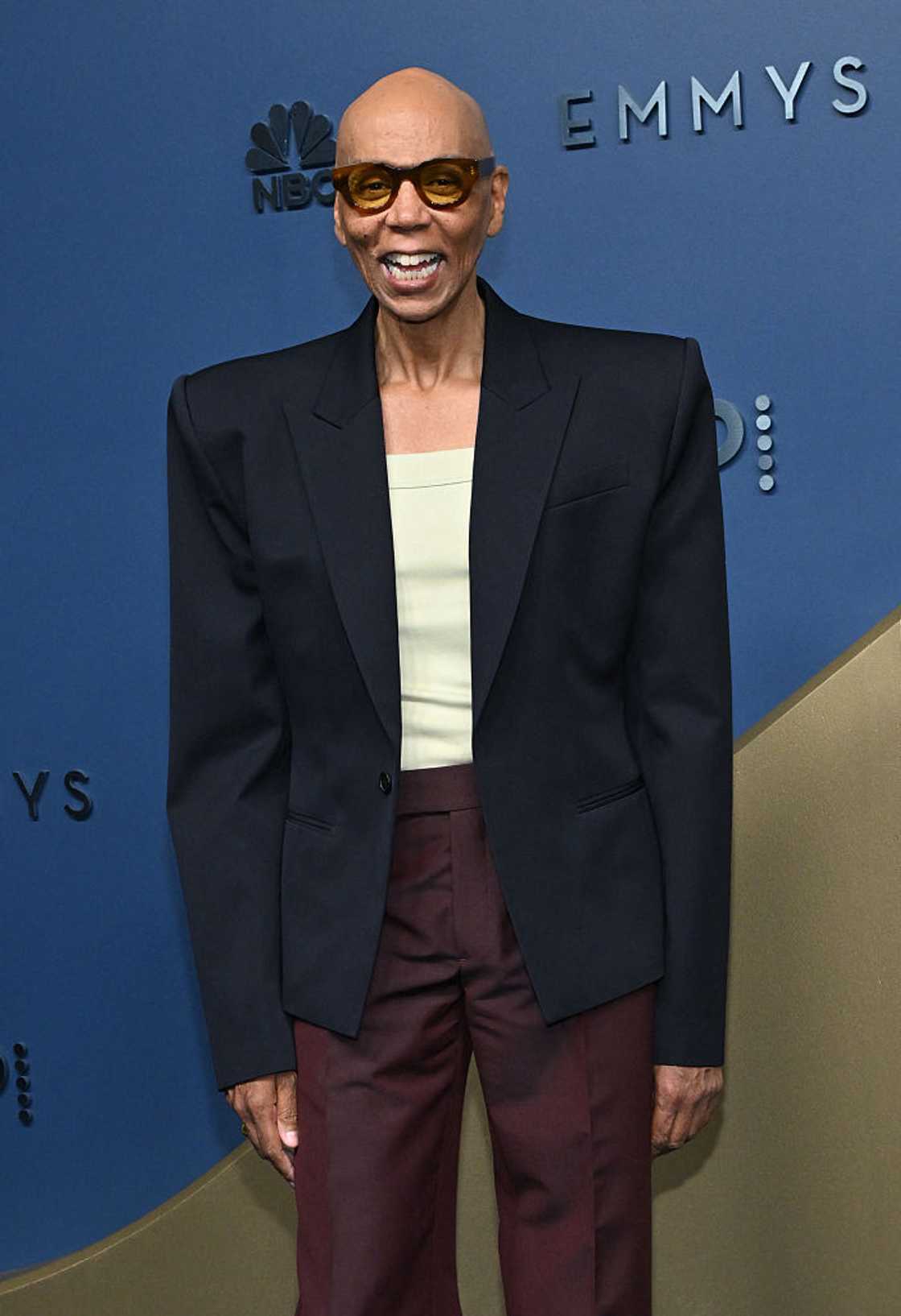 RuPaul in tinted glasses, a black blazer and maroon trousers posing in front of an Emmys backdrop. RuPaul in tinted glasses, a black blazer and maroon trousers posing in front of an Emmys backdrop.