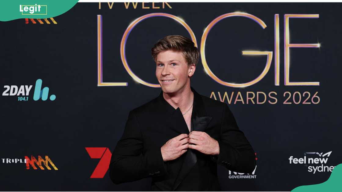 A man in a black suit adjusts his jacket lapels while smiling in front of the Logie Awards 2026 step-and-repeat. A man in a black suit adjusts his jacket lapels while smiling in front of the Logie Awards 2026 step-and-repeat.