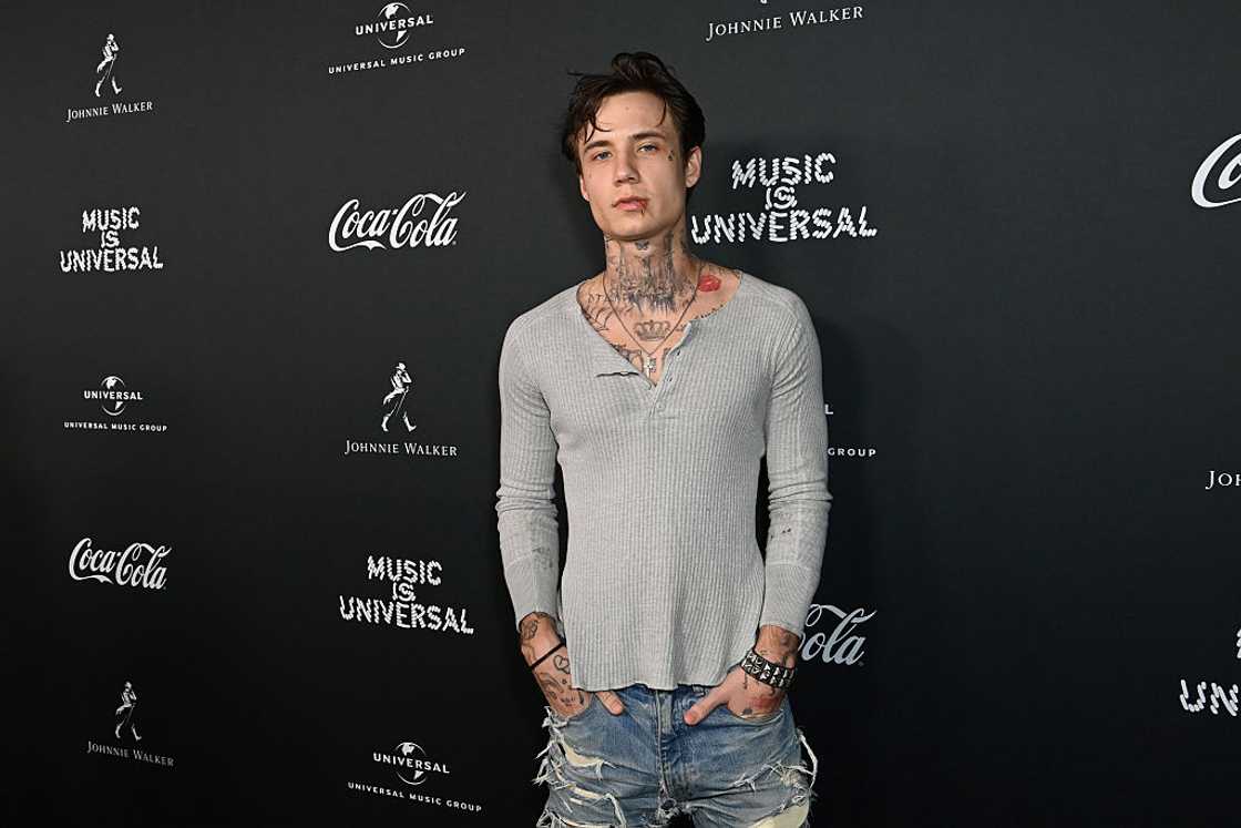 A young man with dark hair and neck tattoos poses in a grey henley. A young man with dark hair and neck tattoos poses in a grey henley.