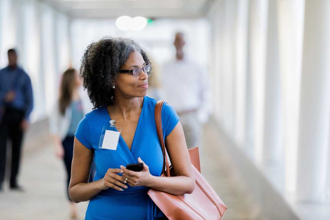 Woman in a blue dress stands in a bright corridor holding a phone and tote bag. Woman in a blue dress stands in a bright corridor holding a phone and tote bag.