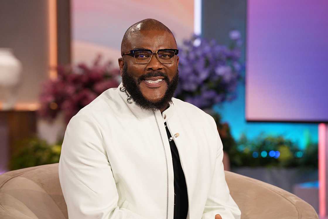 Tyler Perry wearing glasses and a white jacket over a black top, smiling while seated on a talk show set.