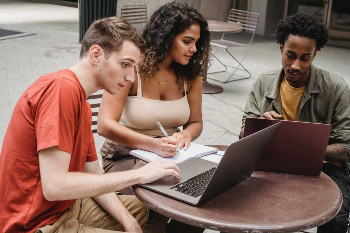 A lady is writing ideas, and two guys are working on their laptops in the cafeteria