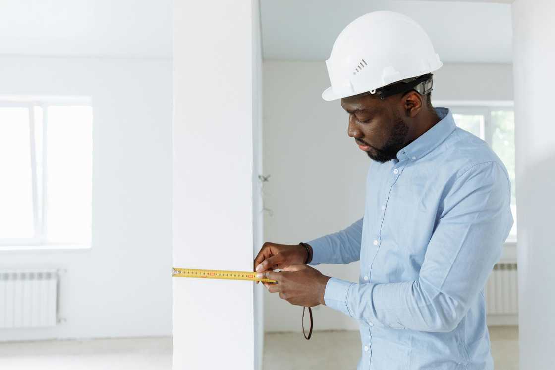 An engineer in a blue long-sleeved shirt and white helmet is using a measuring tape on a house pillar An engineer in a blue long-sleeved shirt and white helmet is using a measuring tape on a house pillar