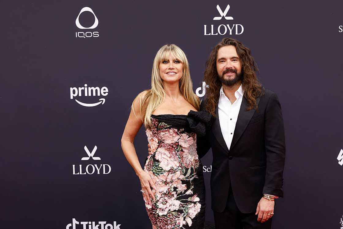 Heidi in a pink and black floral sequined off-shoulder gown stands beside a man in a black tuxedo with a white shirt. Heidi in a pink and black floral sequined off-shoulder gown stands beside a man in a black tuxedo with a white shirt.