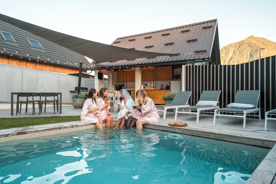 Female friends, clad in silk bathrobes, toasting with champagne at a bachelorette pool party while playfully dipping their legs in the water.