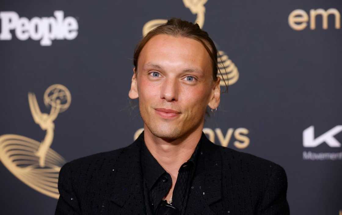 A man with short curly dark hair wears a black jacket over a cream shirt in front of an Apple Original Films backdrop.