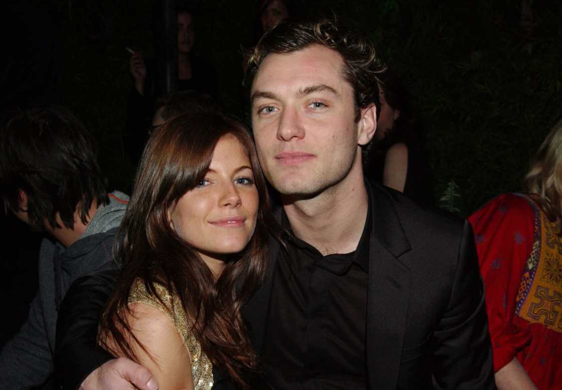 Jude Law in a black suit sits close with a brunette woman in a gold top at a dimly lit gathering. Jude Law in a black suit sits close with a brunette woman in a gold top at a dimly lit gathering.