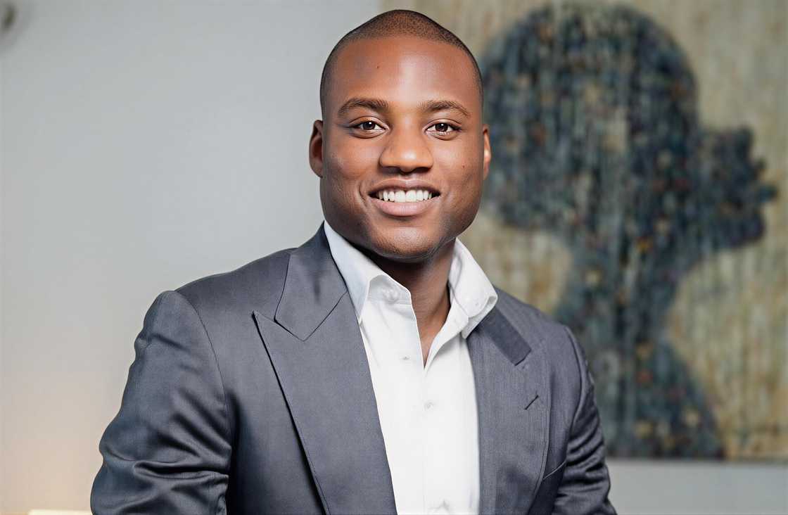 Ladi Delano poses sitting on a couch, wearing a gray suit and white shirt