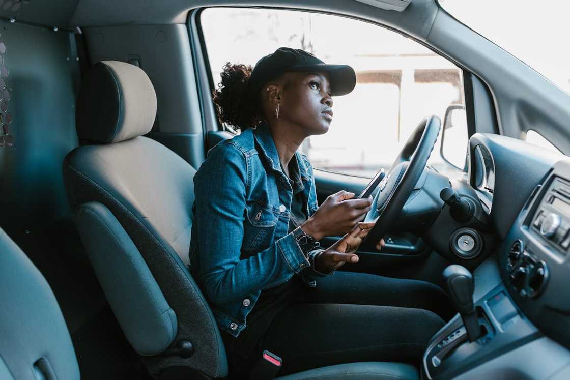 A woman sits in a van holding her phone, looking ahead thoughtfully.