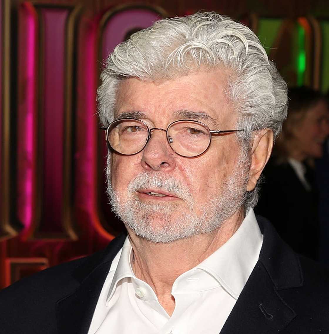 George Lucas wearing round glasses and a black blazer over a white shirt, standing at an event with colorful lights behind him. George Lucas wearing round glasses and a black blazer over a white shirt, standing at an event with colorful lights behind him.